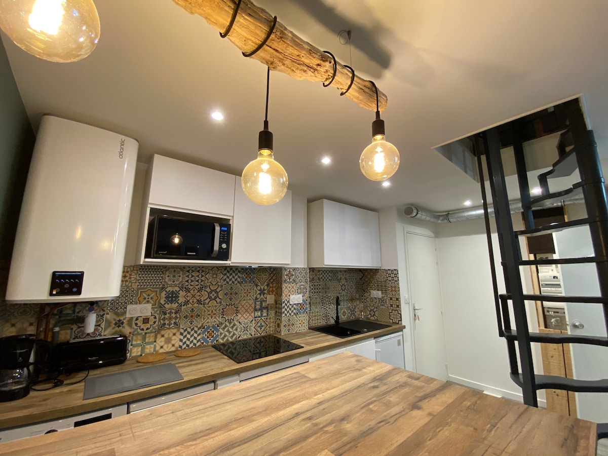 The kitchen area features modern appliances, including a refrigerator, microwave, and induction cooktop. The countertop is complemented by a mosaic tile backsplash. Above, three pendant lights provide illumination, and a staircase leads to the mezzanine.
