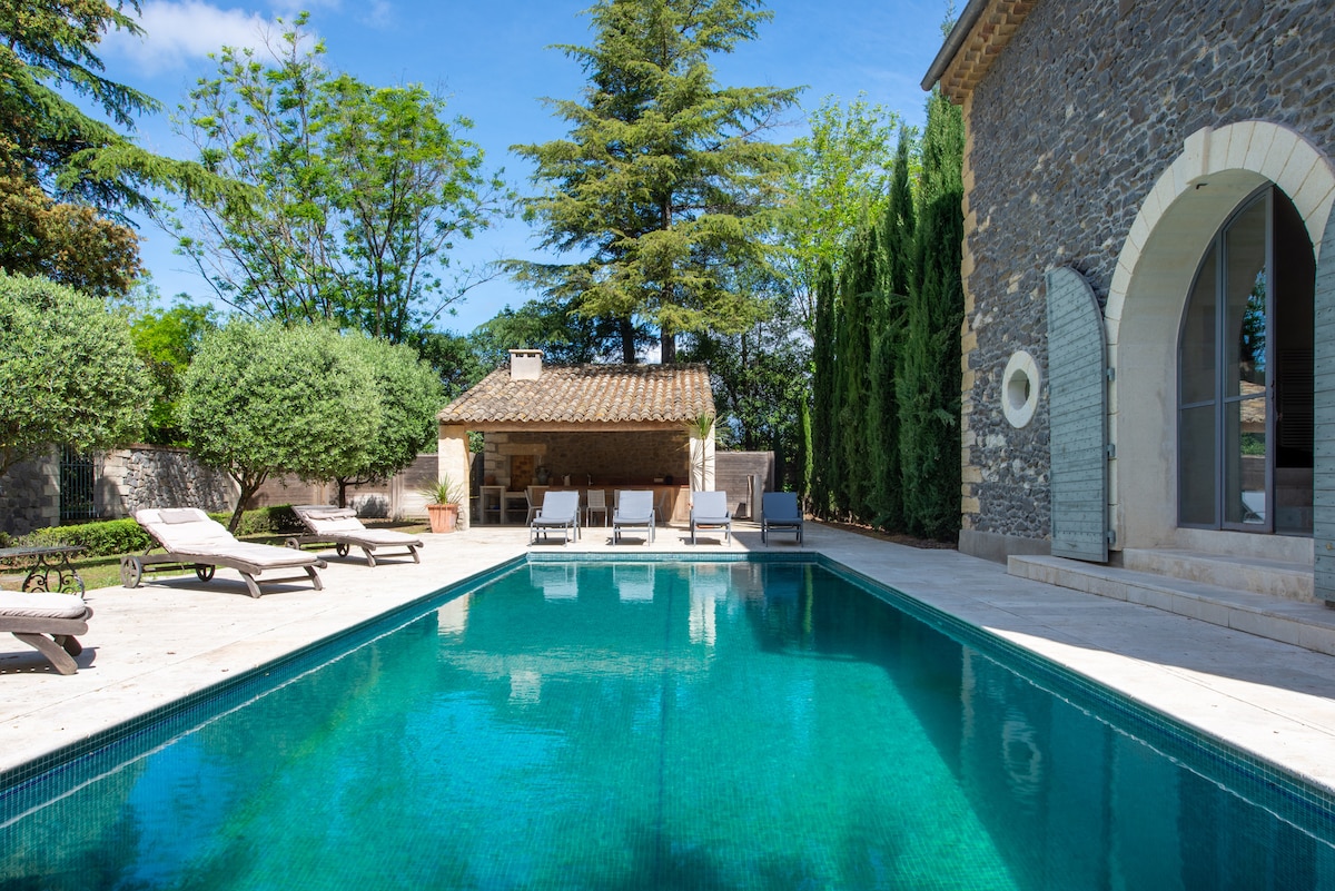 A serene pool area is surrounded by lush greenery and mature trees. Several lounge chairs are placed around the pool, complementing the inviting turquoise water. A rustic outbuilding with a tiled roof is visible in the background, adding charm to the outdoor space.