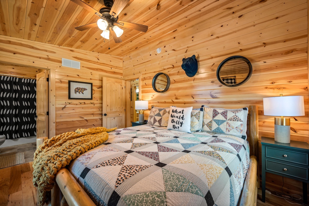 A cozy bedroom features a spacious bed adorned with a patterned quilt and decorative pillows. Two round mirrors hang above nightstands with lamps on either side. Wood paneling lines the walls, complemented by a light fixture with multiple bulbs.