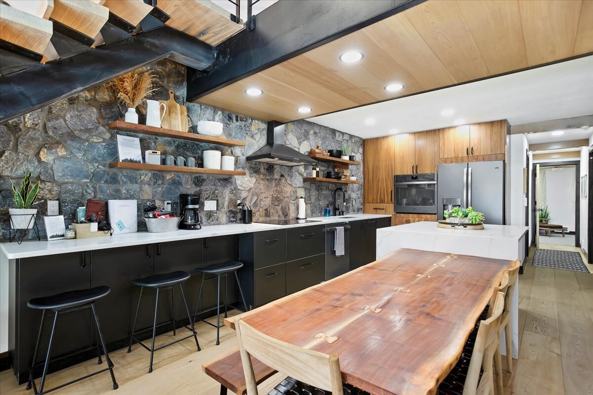 A modern kitchen is featured with a stylish stone accent wall. A long wooden dining table is surrounded by black chairs, and kitchen appliances are neatly arranged along the counter. Natural light enhances the warm tones of the cabinetry and decor.