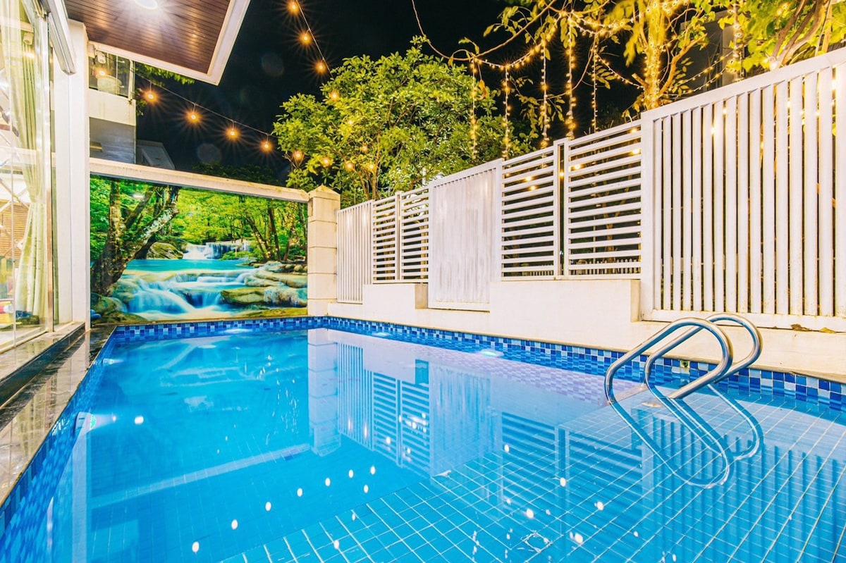 The outdoor pool area features a clear blue swimming pool framed by a white fence. Soft lighting from hanging bulbs creates a warm ambiance. A serene backdrop of a natural scene is visible on the adjacent wall, enhancing the tranquil setting.