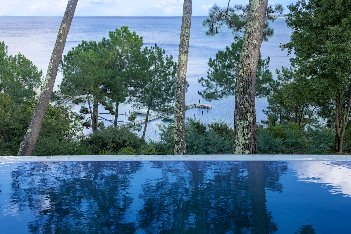 An infinity pool reflects the surrounding trees and sky, creating a seamless connection with the tranquil lake below. A wooden pier extends into the water, framed by lush greenery. The scene emphasizes a calm and serene landscape, inviting relaxation.