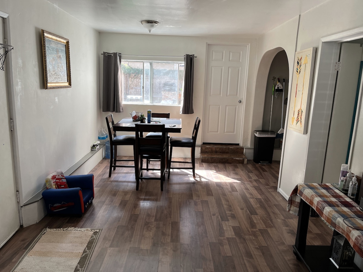 A bright and spacious common area is seen, featuring a dining table with chairs positioned near a large window. Light-colored walls and hardwood flooring contribute to an open feel. A doorway leads to another room, while a small rug and some items are visible in the lower left corner.