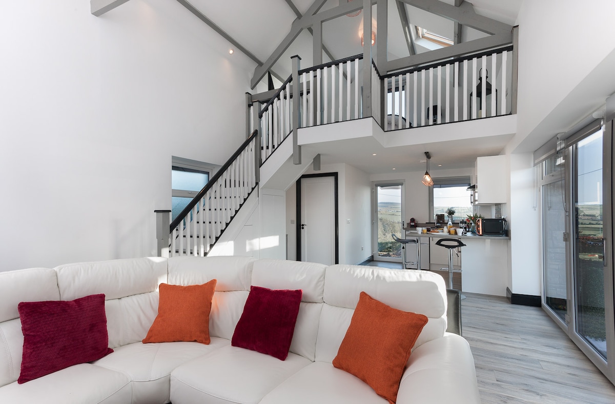 A cozy living area features a white sectional sofa adorned with orange and red throw pillows. The open-plan layout offers views of a modern kitchen in the background and a staircase leading to the mezzanine level, surrounded by vaulted ceilings and natural light.