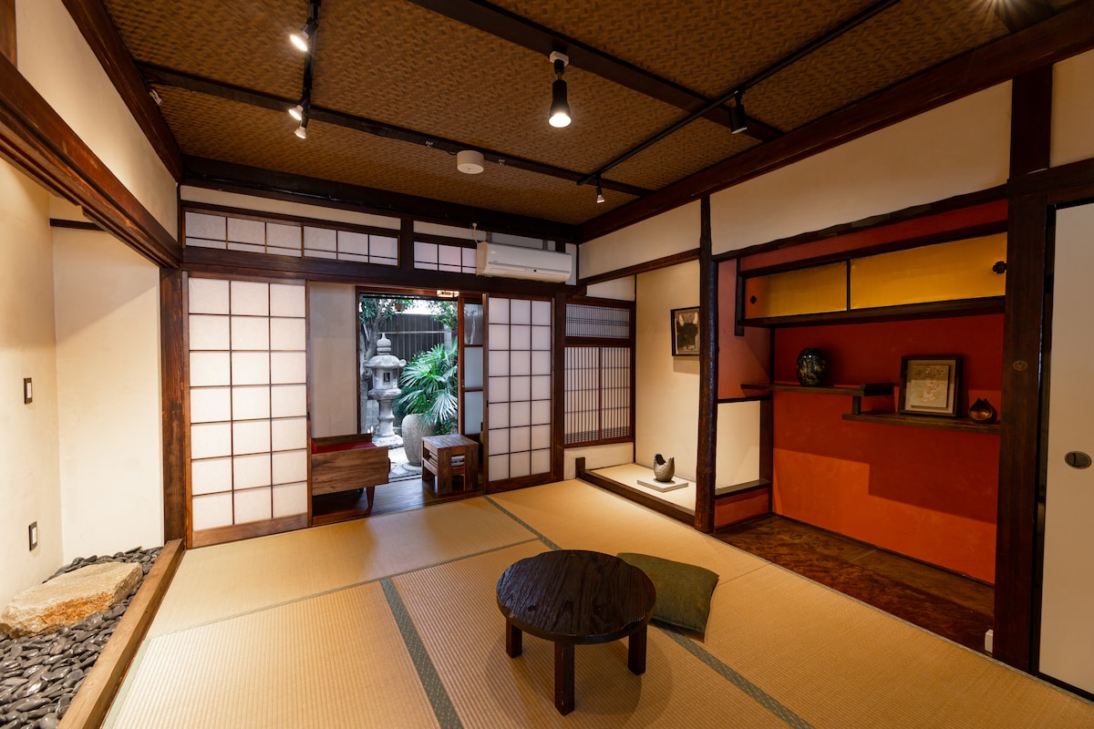 A traditional Japanese room features tatami flooring and sliding shoji doors leading to a garden view. Natural light illuminates the space, where a low wooden table is placed centrally on the floor. The walls are adorned with warm colors and decorative elements, creating a calming atmosphere.