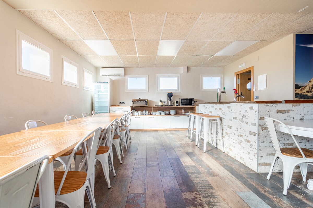 A spacious dining area features a long wooden table surrounded by several white chairs. Light-colored cabinetry and counter space are visible in the background, complemented by appliances. The room is illuminated by natural light flowing through multiple small windows.