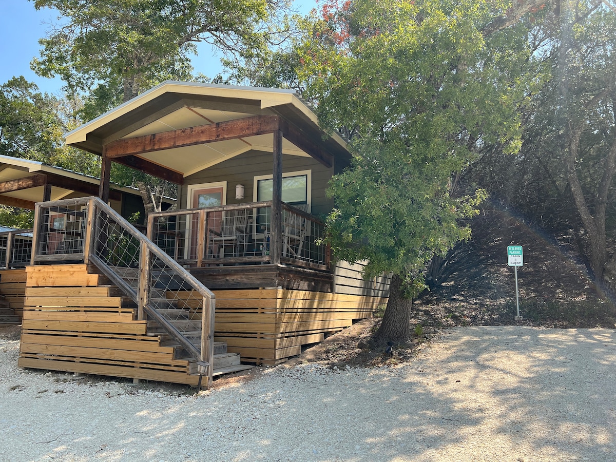 A cabin with a welcoming porch is elevated on a wooden platform, surrounded by natural landscaping. Sturdy steps lead to the entry, and the surrounding area features gravel paths and mature trees, enhancing the sense of privacy and connection to nature.