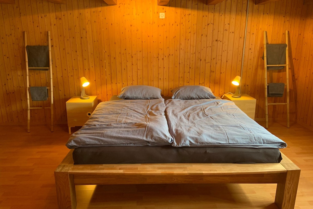 A cozy sleeping area features a spacious bed with gray bedding, situated on a wooden frame. Warm wood paneling adorns the walls, complemented by soft lighting from bedside lamps on wooden nightstands. Two folded towels are displayed on decorative ladders nearby.