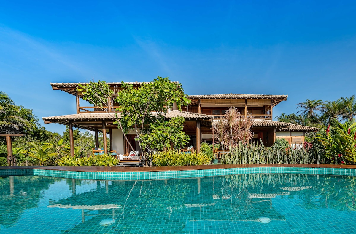 A spacious wooden house is set against a clear blue sky, surrounded by lush tropical foliage. An infinity-edge saltwater pool reflects the vibrant greenery and provides a serene atmosphere. Sun loungers are visible on the deck, inviting relaxation.