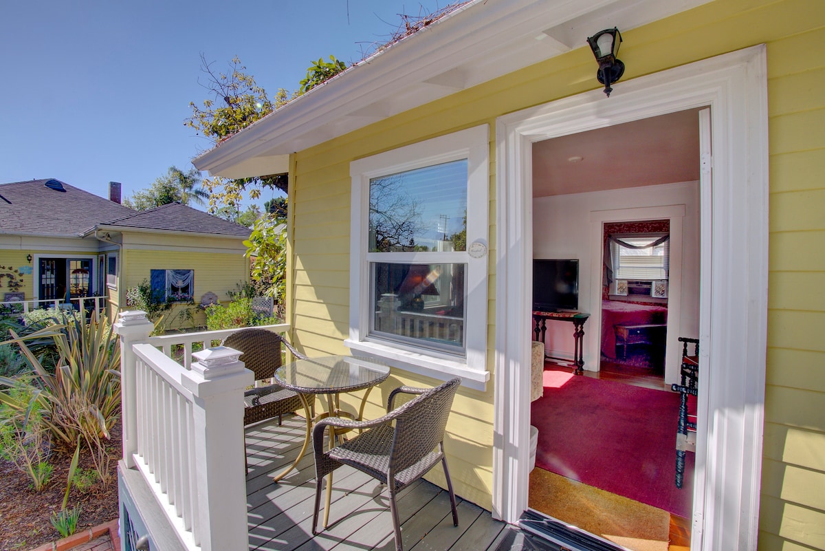 An exterior view of a charming cottage features a small deck with two woven chairs and a table. A bright yellow façade is complemented by white trim. Through the open door, a cozy interior space is visible, with a television and seating area in view.