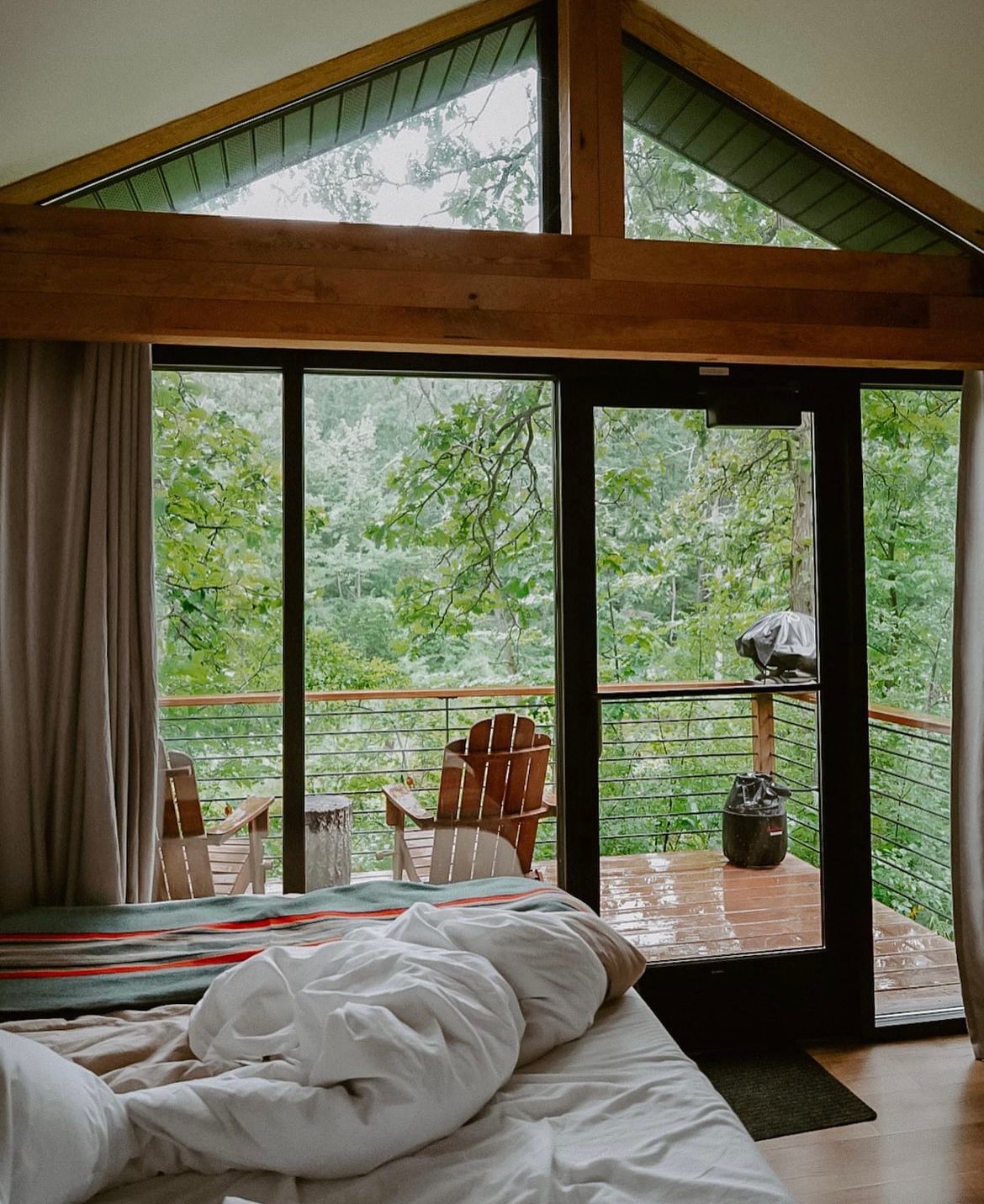 A spacious sleeping area features a rumpled white duvet on a large bed. Floor-to-ceiling glass doors provide views of the lush greenery outside, leading to a private deck with wooden chairs and a small table. Natural light floods the room, accentuating wooden beams.