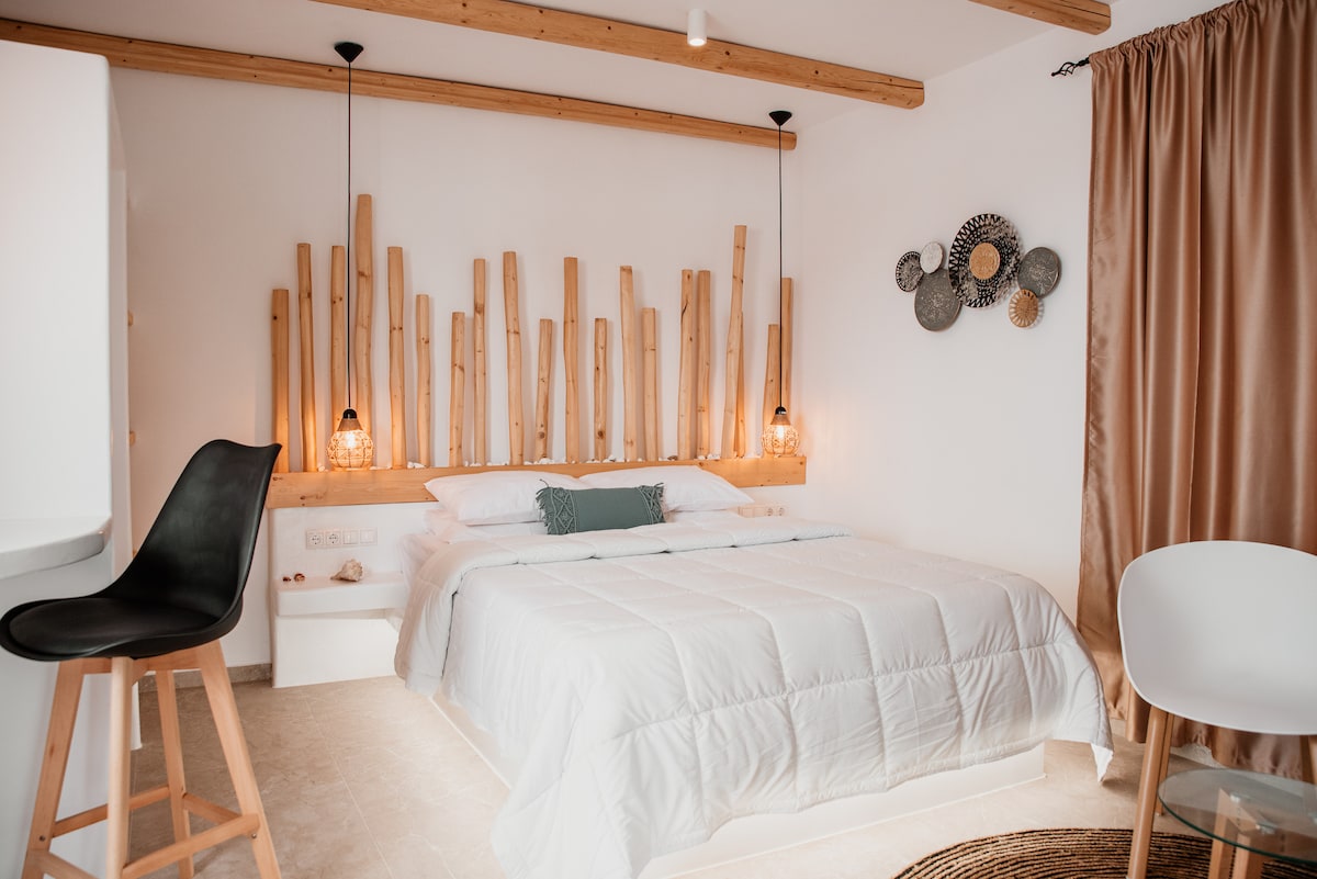 A bright sleeping area is centered around a spacious bed framed by natural wood accents. Pendant lights provide soft illumination, while artwork adds texture to the walls. A modern chair and a minimalist table complement the open layout, enhancing the serene environment.