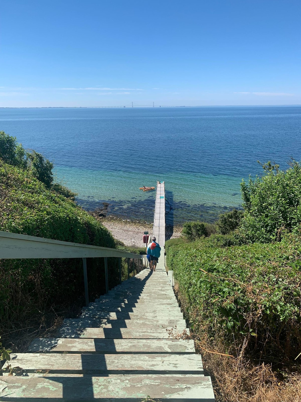 A wooden staircase leads down towards a tranquil beach, bordered by lush greenery on either side. The clear water reveals a gradient from deep blue to light turquoise, with a wooden pier extending into the ocean. The serene landscape under a clear blue sky creates a peaceful atmosphere.