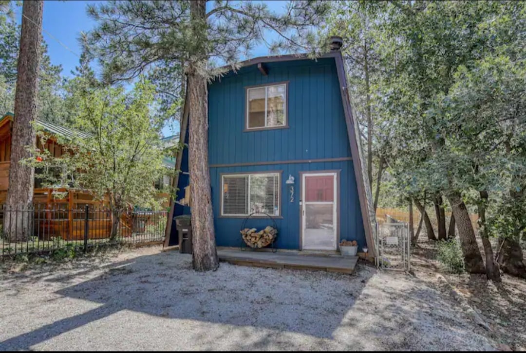 A blue cabin features an a-frame design surrounded by tall trees. A welcoming entrance is highlighted by a wooden porch, while a stacked log pile sits nearby. The gravel yard provides an open space, enhancing the cabin's connection to nature.