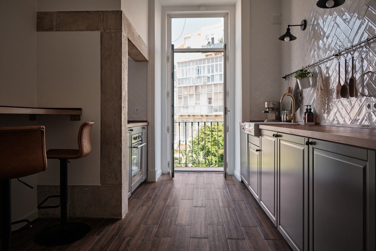 A modern kitchen features sleek cabinetry and natural wood countertops. Large windows bring in natural light, revealing a view of the street outside. A circular table with two chairs is positioned next to the kitchen area, and various utensils and kitchen tools are arranged neatly along the wall.