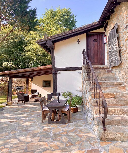 A stone staircase leads to an upper-level entrance, flanked by a wooden porch area. A dining table with four chairs is positioned on the stone patio, surrounded by greenery. The exterior displays a rustic charm, blending seamlessly with the natural surroundings.