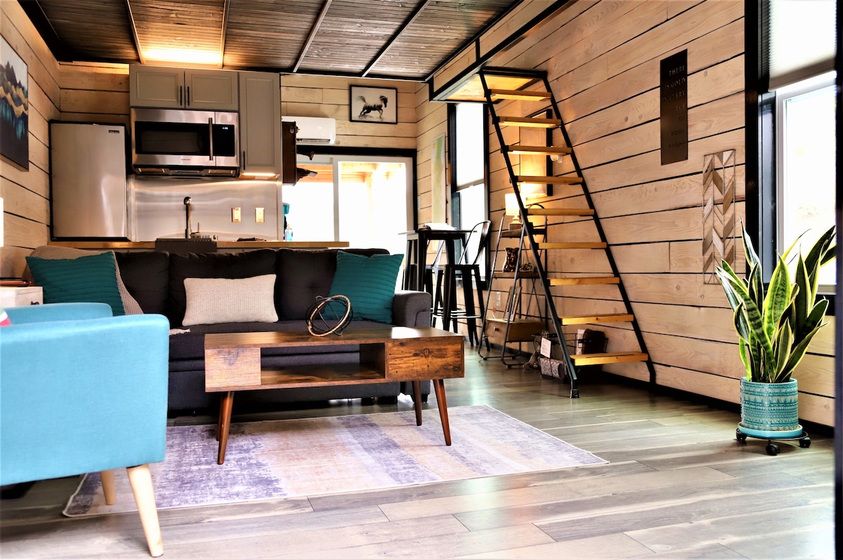 The interior space showcases modern design elements, featuring a cozy seating area with a sectional sofa and a wooden coffee table. A bright kitchen area is visible, equipped with stainless steel appliances. Stairs lead to an upper loft, illuminated by natural light from large windows.