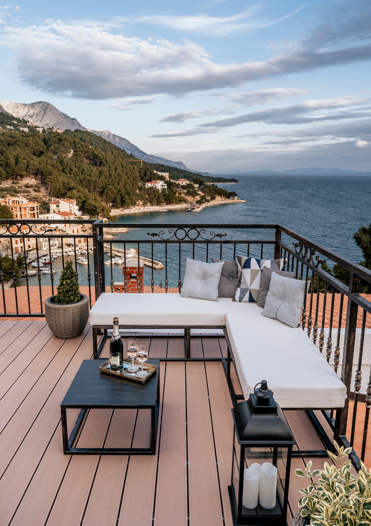 An outdoor seating area features a modern sectional sofa adorned with cushions. A small table holds glasses and a bottle, with a lantern positioned beside it. The balcony offers a stunning view of the coastline and mountainous landscape under a partly cloudy sky.