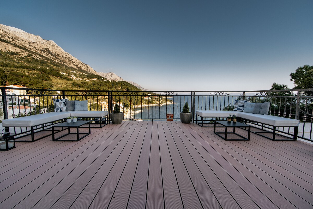 A spacious covered terrace features comfortable seating areas divided by a clear railing, offering views of the surrounding landscape. Several potted plants are positioned alongside the seating, creating a welcoming atmosphere. The deck has a light wood finish, complementing the serene surroundings.