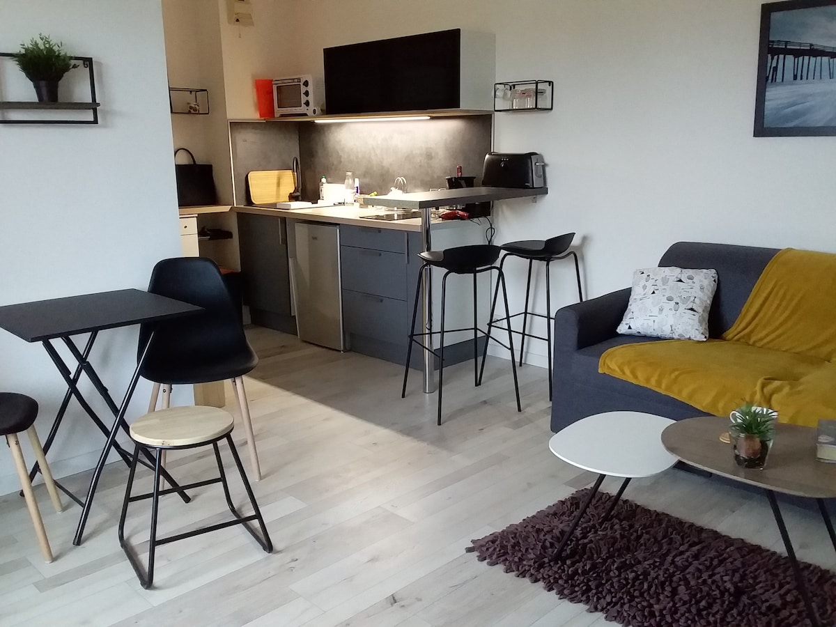 An open living space features a kitchen area with modern appliances, including a microwave. A dining table with four chairs is positioned next to a cozy seating area, which includes a sofa and various side tables. Natural light brightens the room.