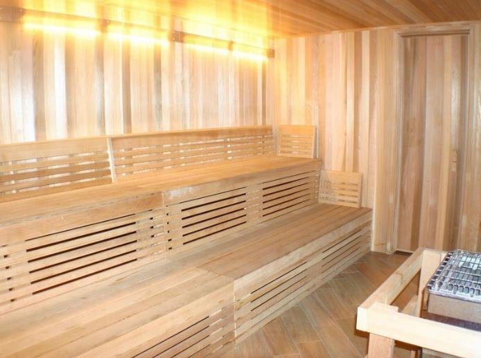 A wooden sauna interior is showcased, featuring bench seating made of slatted wood. The warm tones of the wood create a cozy environment. Soft lighting is present along the upper wall, enhancing the serene ambiance. A small corner is designated for heating elements.
