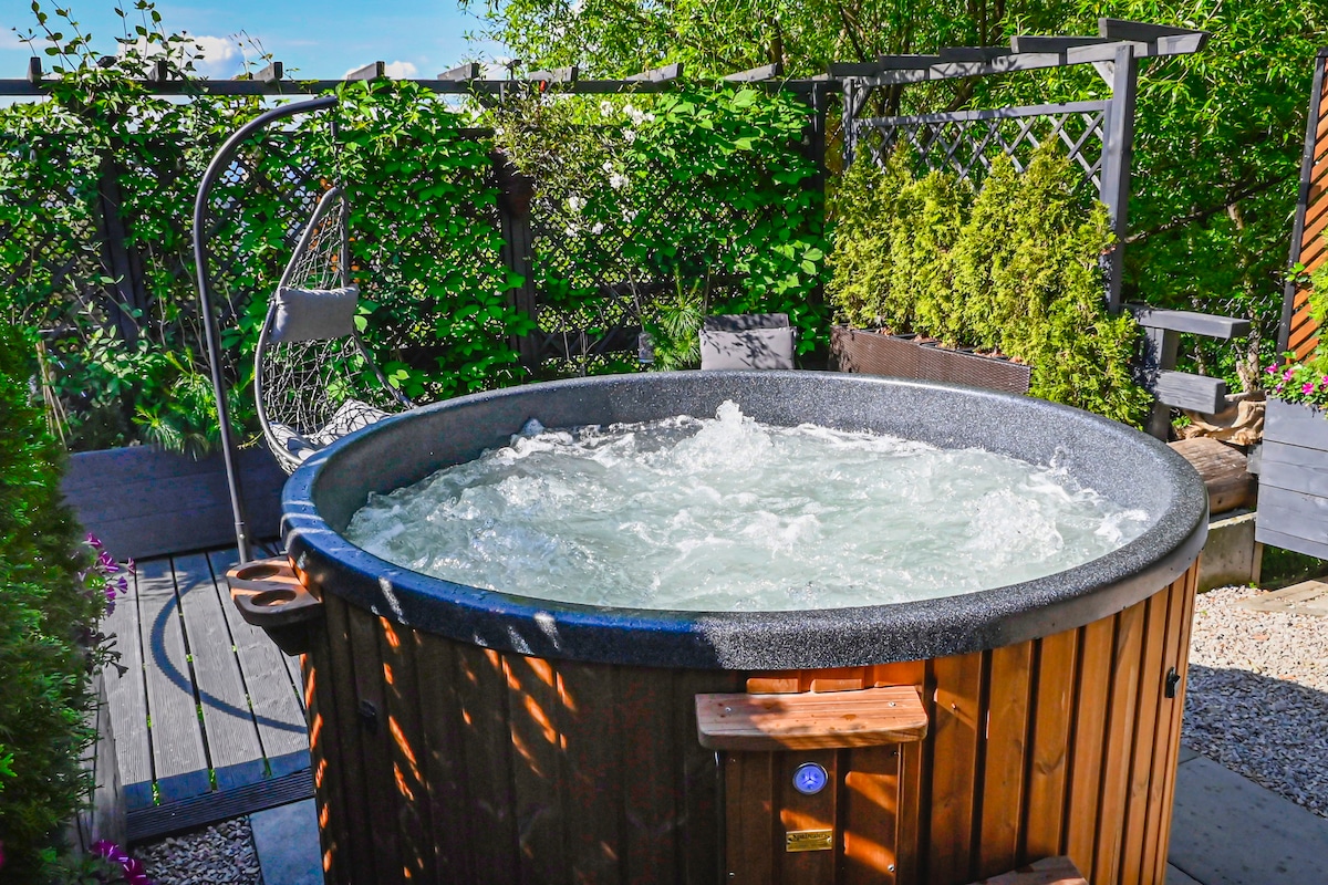 An outdoor jacuzzi is situated in a private corner, surrounded by lush greenery and trellises. The bubbling water is inviting and illuminated by natural sunlight, creating a serene relaxation space.