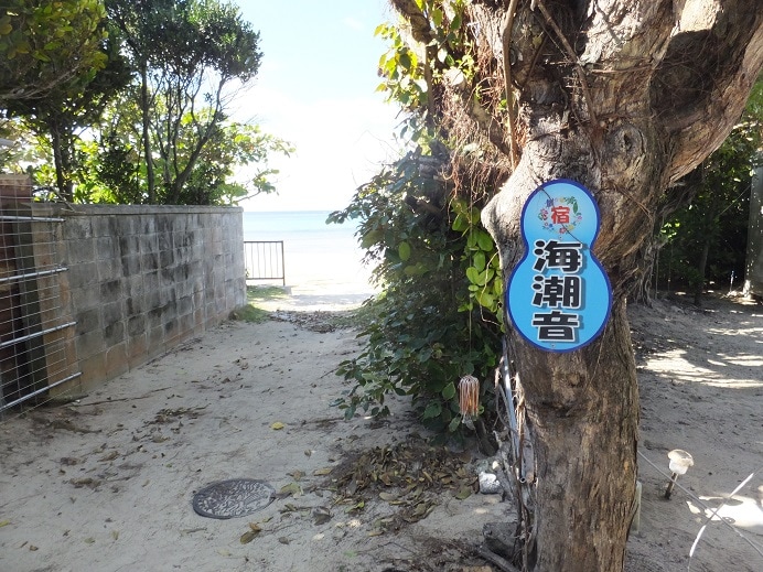 A tree stands at the property edge, adorned with a sign featuring the name of the listing. Behind the tree, a sandy pathway leads to a view of the sea, bordered by greenery and a stone wall.