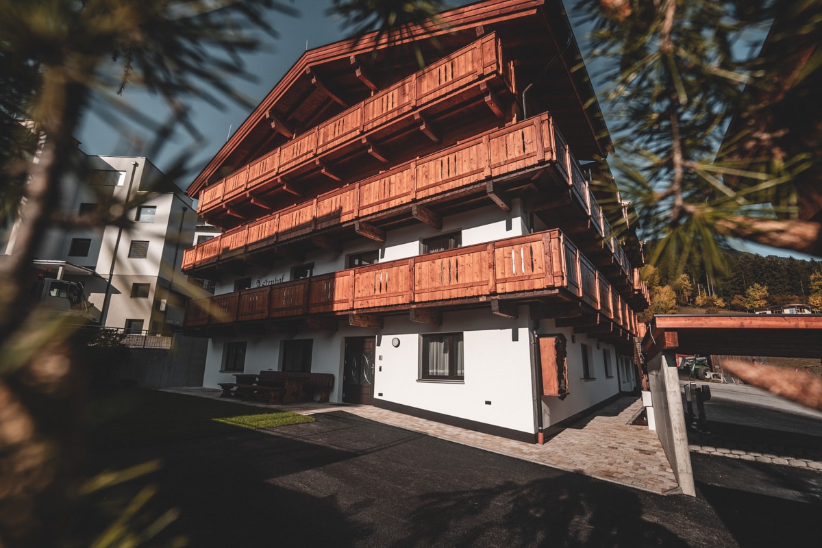 The three-story building features striking wooden balconies with a rustic exterior. The entrance is framed by greenery, and the surrounding area includes paved paths leading to the entrance. Ample outdoor space is visible, enhancing the inviting atmosphere of the property.