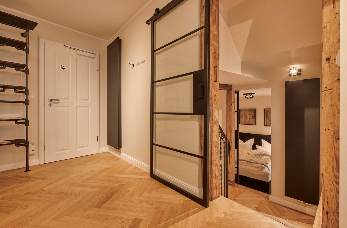 A welcoming entryway features a blend of wooden and modern design elements. Light wood parquet flooring complements the light-colored walls. Glass doors provide a view into the cozy sleeping area, while a stylish black radiator and decorative metal accents enhance the overall aesthetic.