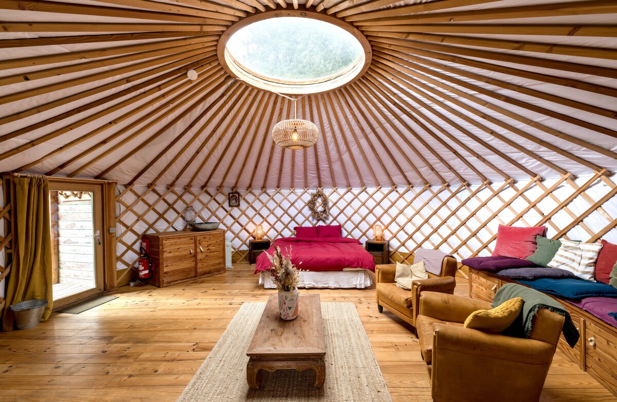 The interior of the yurt features a circular skylight that allows natural light to flood the space. Wooden beams create an open and airy atmosphere. A double bed is surrounded by colorful cushions, while a cozy seating area includes two armchairs and a wooden coffee table.