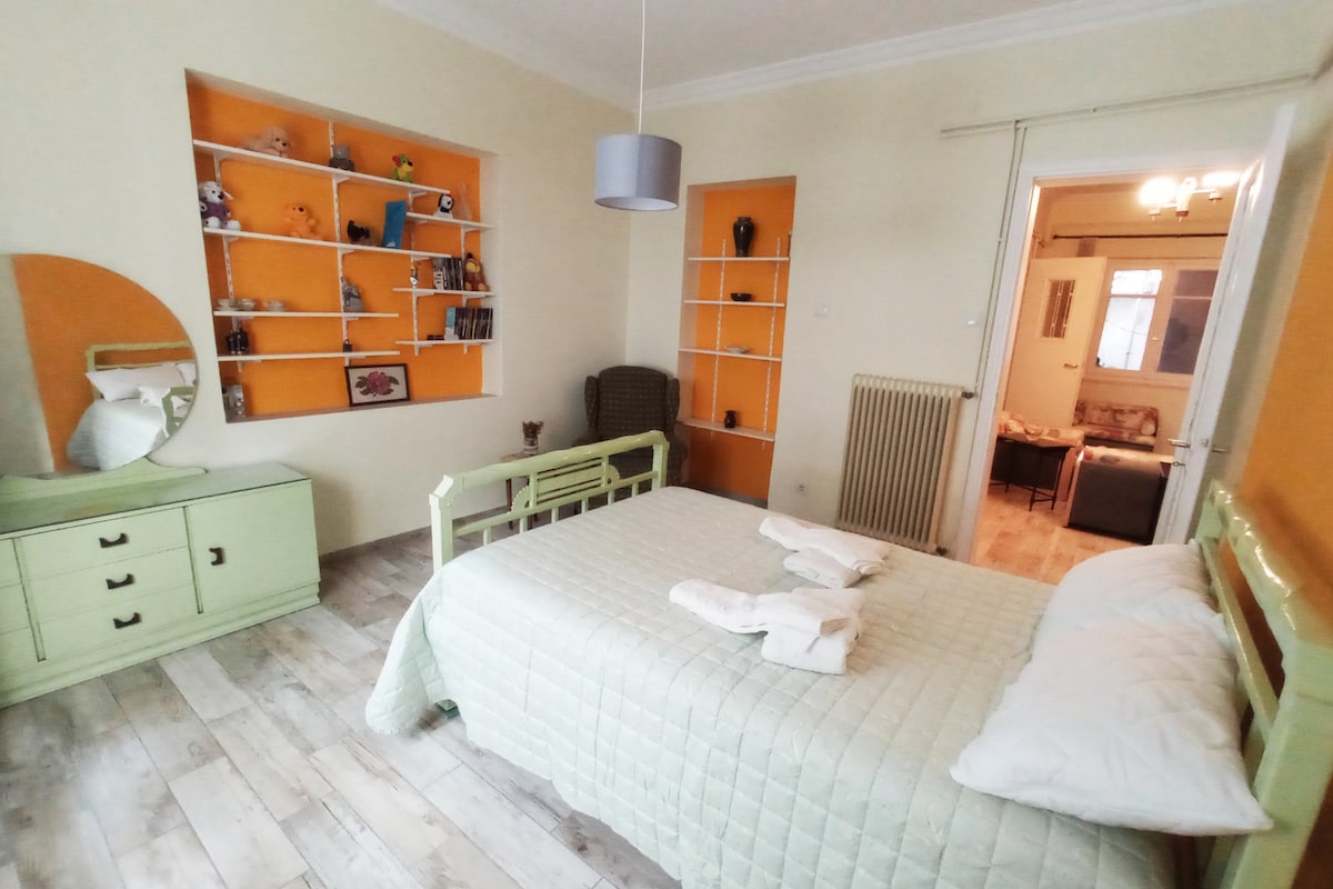 A spacious bedroom features a light green bedframe and pale quilt. Adjacent walls showcase bright orange accents. A round mirror and a dresser are present, while shelves host various decorative items. A doorway leads to a living area, inviting natural light into the room.