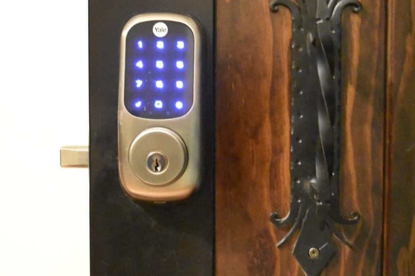 A digital door lock with a keypad is displayed, featuring illuminated blue buttons. The lock is mounted on a wooden door that showcases an ornate black handle, offering a classic contrast with the technological design.