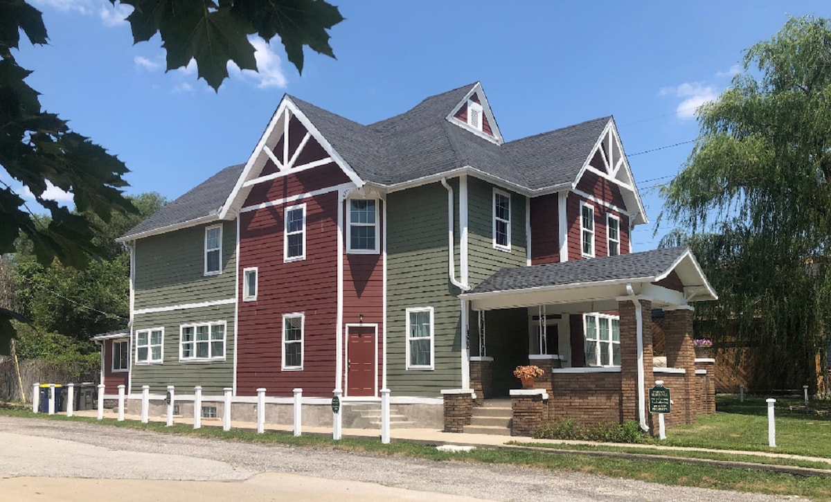 The exterior of the house is presented, featuring a mix of green and red siding with white architectural accents. A welcoming front porch is visible, flanked by landscaped areas. The structure has multiple windows and distinct sloped roofs, contributing to its spacious appearance.