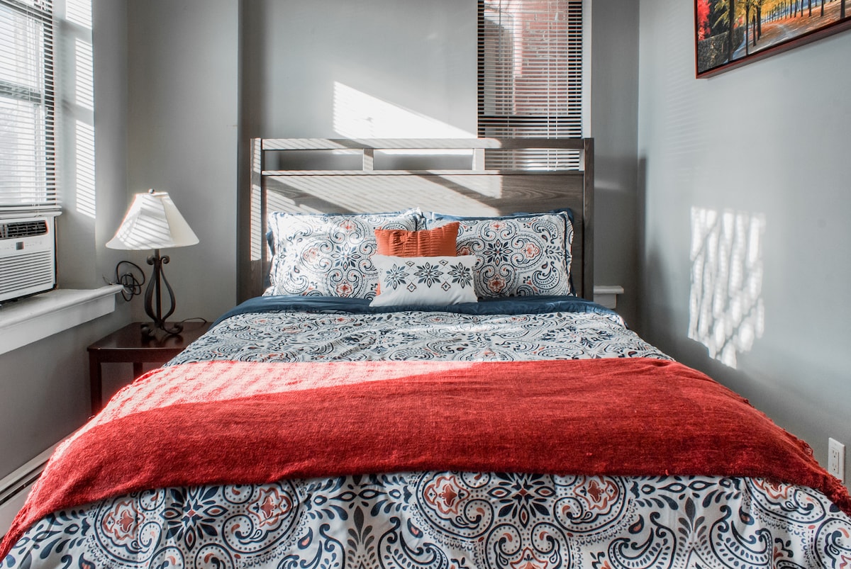 A cozy bedroom features a spacious bed adorned with patterned bedding and a solid red throw blanket. Natural light streams through partially open blinds, illuminating the room. A bedside lamp and wall art enhance the inviting atmosphere.