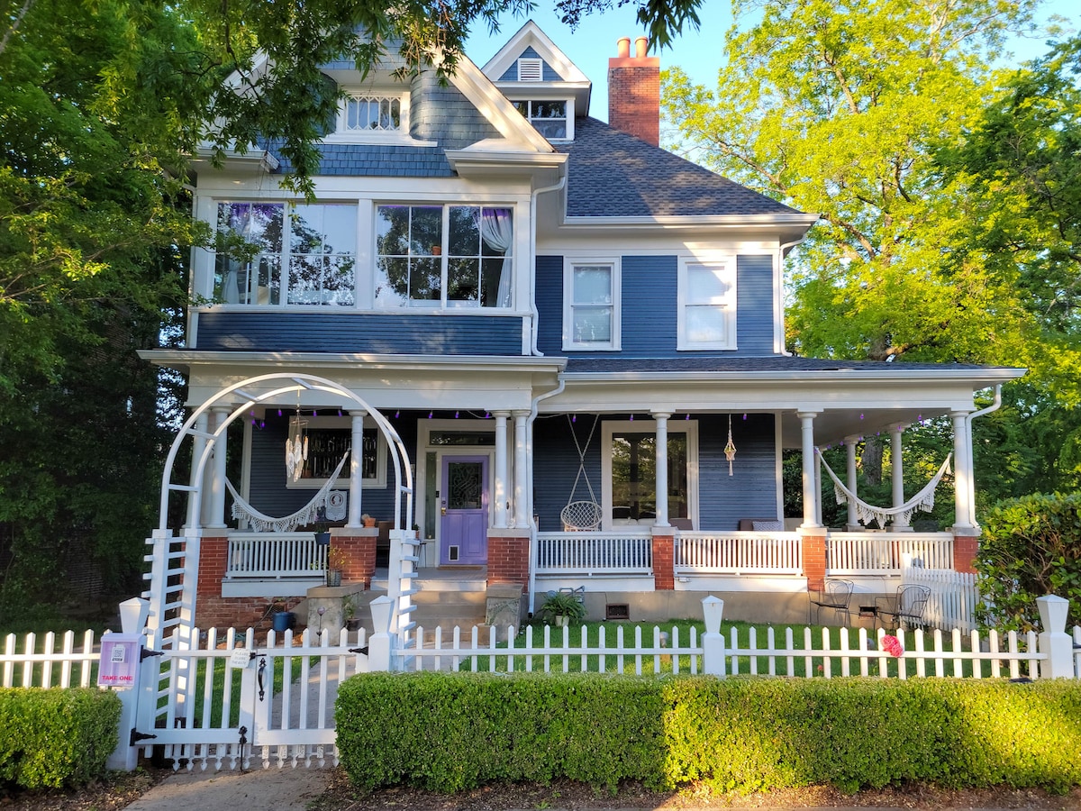 A historic mansion with blue siding and a large porch sits amid lush green trees. The front yard is bordered by a white picket fence, with two hammocks gently swaying on the porch. Large windows illuminate the inviting entrance featuring purple accents.