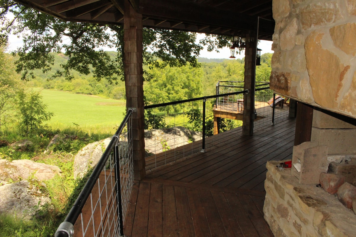 A covered porch extends from the cabin, featuring wooden flooring and steel railings. The view reveals rolling meadows and woods in the distance, with large boulders visible nearby. A fireplace is situated at the edge of the porch, inviting outdoor relaxation.