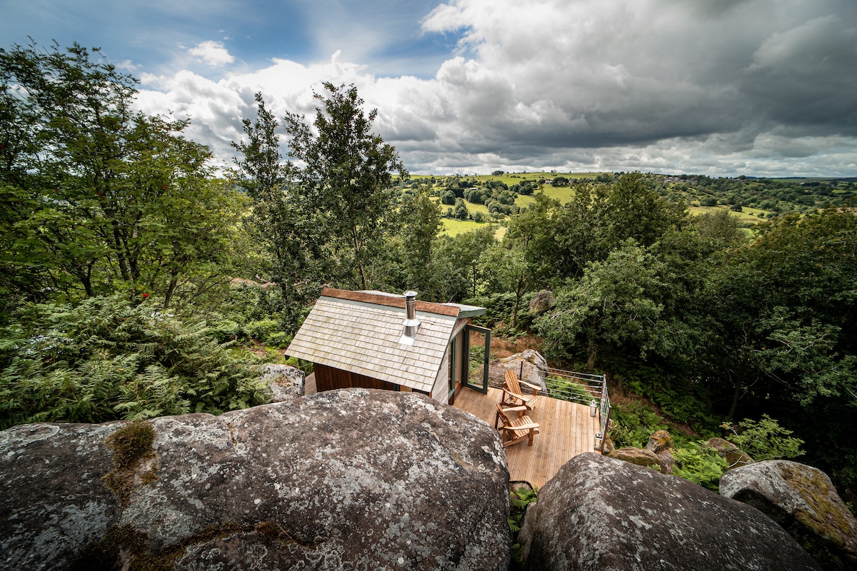 A cabin is perched among large boulders, surrounded by lush greenery and distant rolling hills. The exterior features wooden siding and a sloped roof, complemented by a small deck with chairs. The scene highlights a tranquil, nature-focused setting under a cloudy sky.