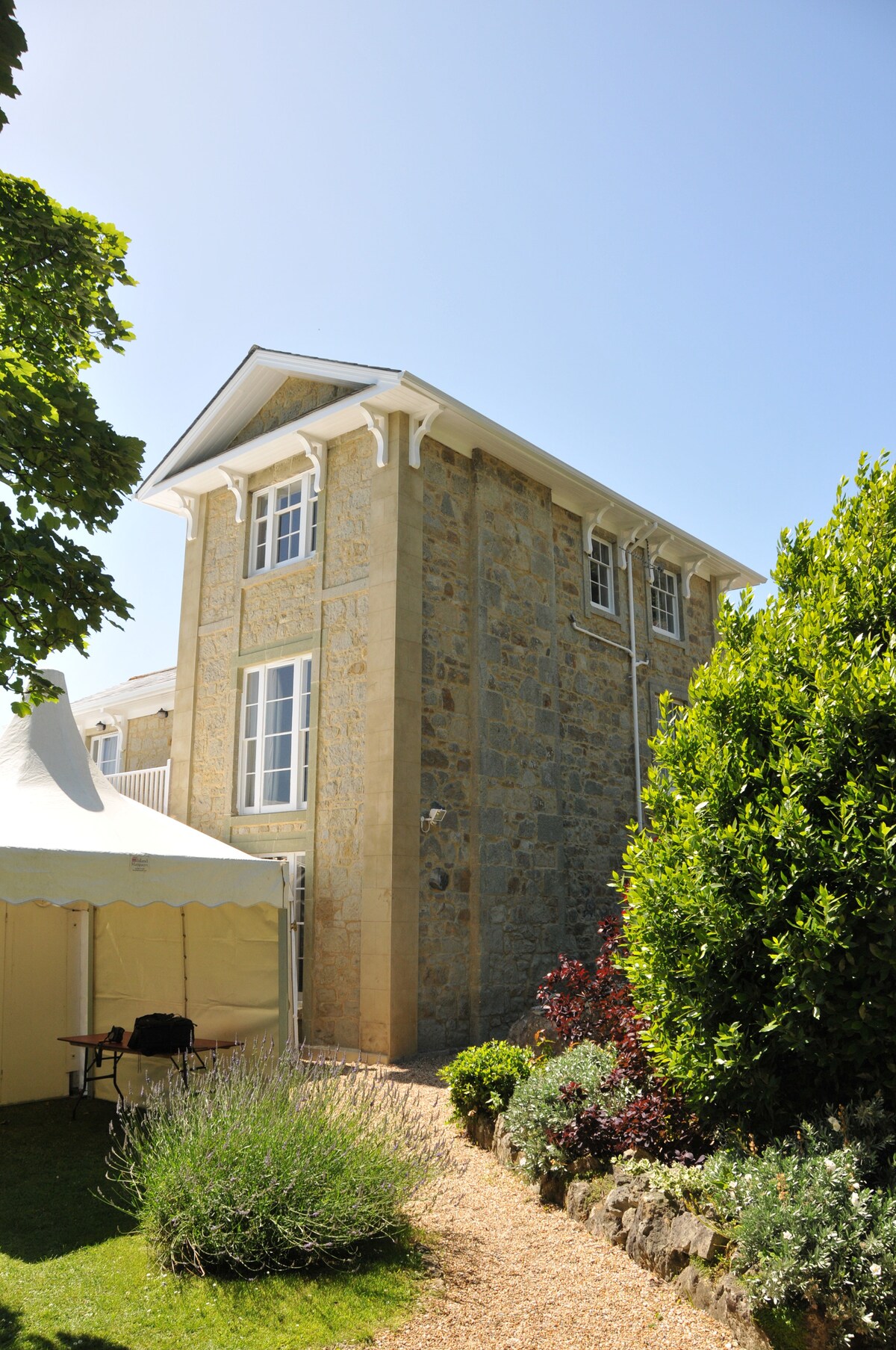 The stone exterior of a multi-story villa is highlighted by large windows and a gabled roof. A pathway lined with greenery leads to the entrance. A white tent stands to the side, adding to the spacious outdoor area.