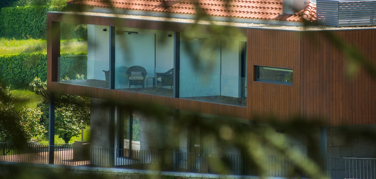 A modern architectural structure is partially visible, featuring large glass panels and a wooden exterior. An outdoor seating area with comfortable chairs is present on the balcony, adorned by a serene green landscape in the background.
