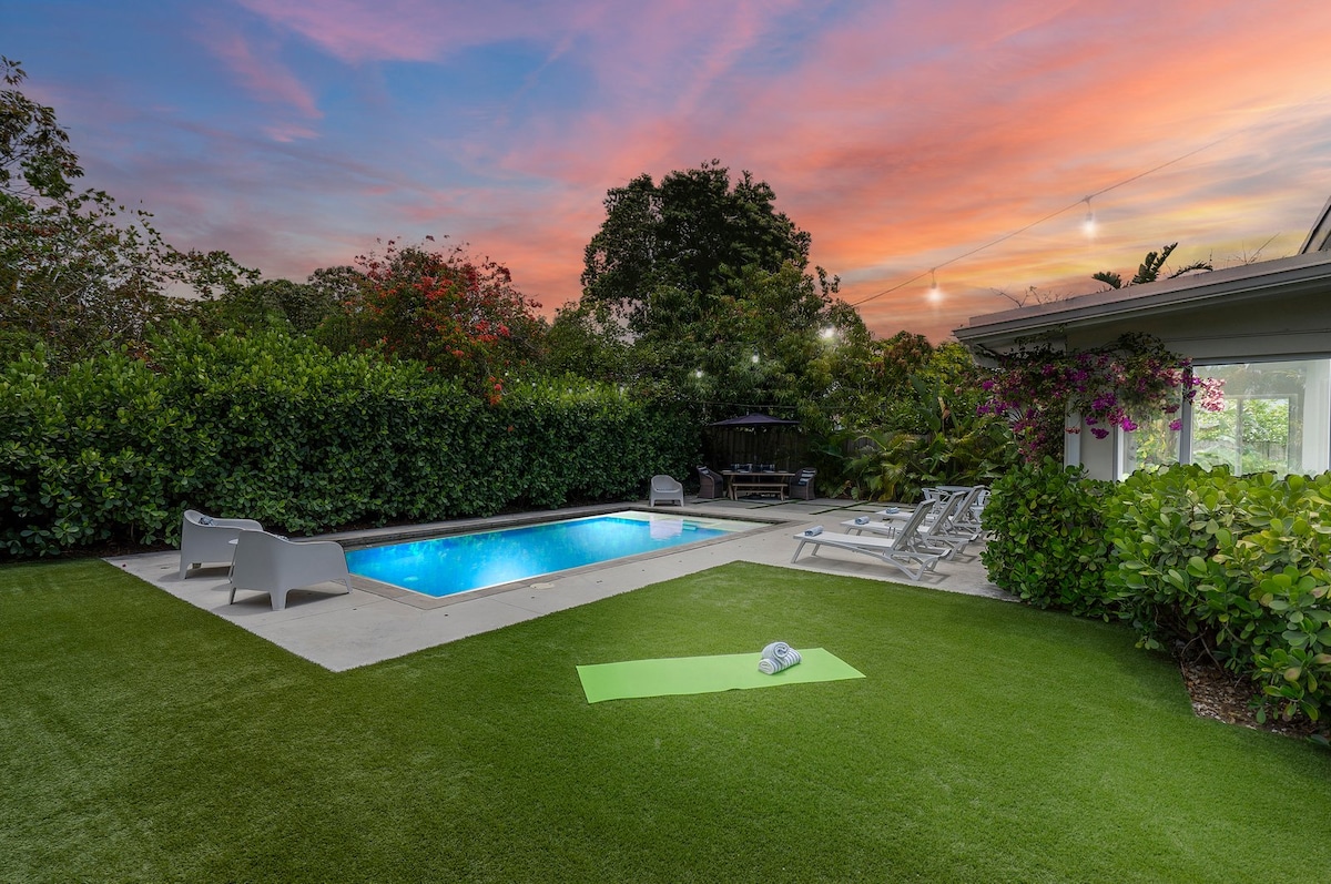 A private heated saltwater pool is framed by a lush tropical garden. Lounge chairs are positioned around the poolside, with a yoga mat resting on the grassy area. The scene is enhanced by a colorful sunset sky, creating a serene outdoor escape.