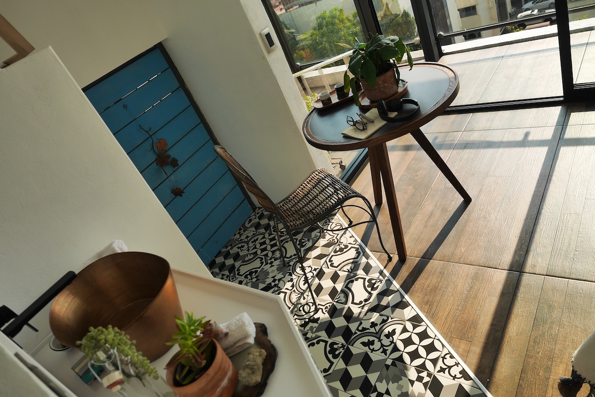 A sunlit corner features a round table with a potted plant and a chair beside it. The floor displays a detailed patterned tile design, contrasting with the wooden plank area. Light filters through the large glass doors, enhancing the warm ambiance.