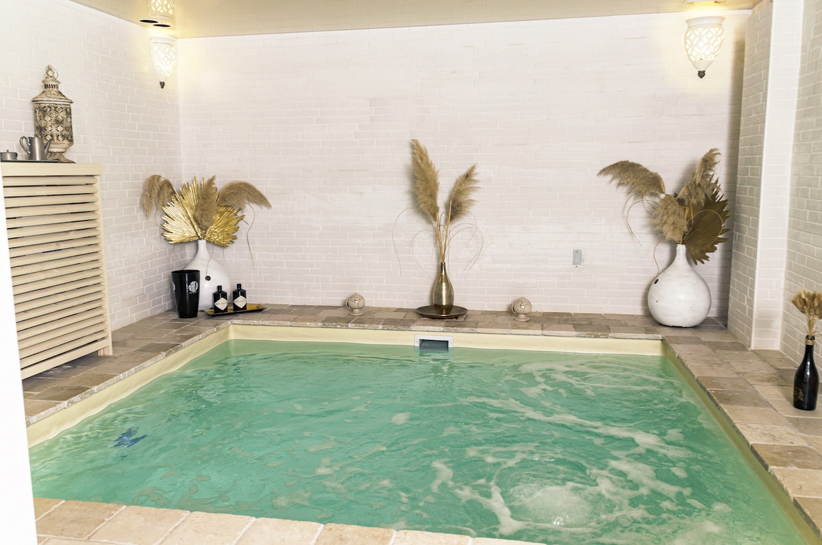 An elegantly designed spa area features a heated jacuzzi surrounded by decorative elements. Soft lighting highlights the natural textures of the space, including plants and tasteful pottery. The tranquil environment invites relaxation and well-being.
