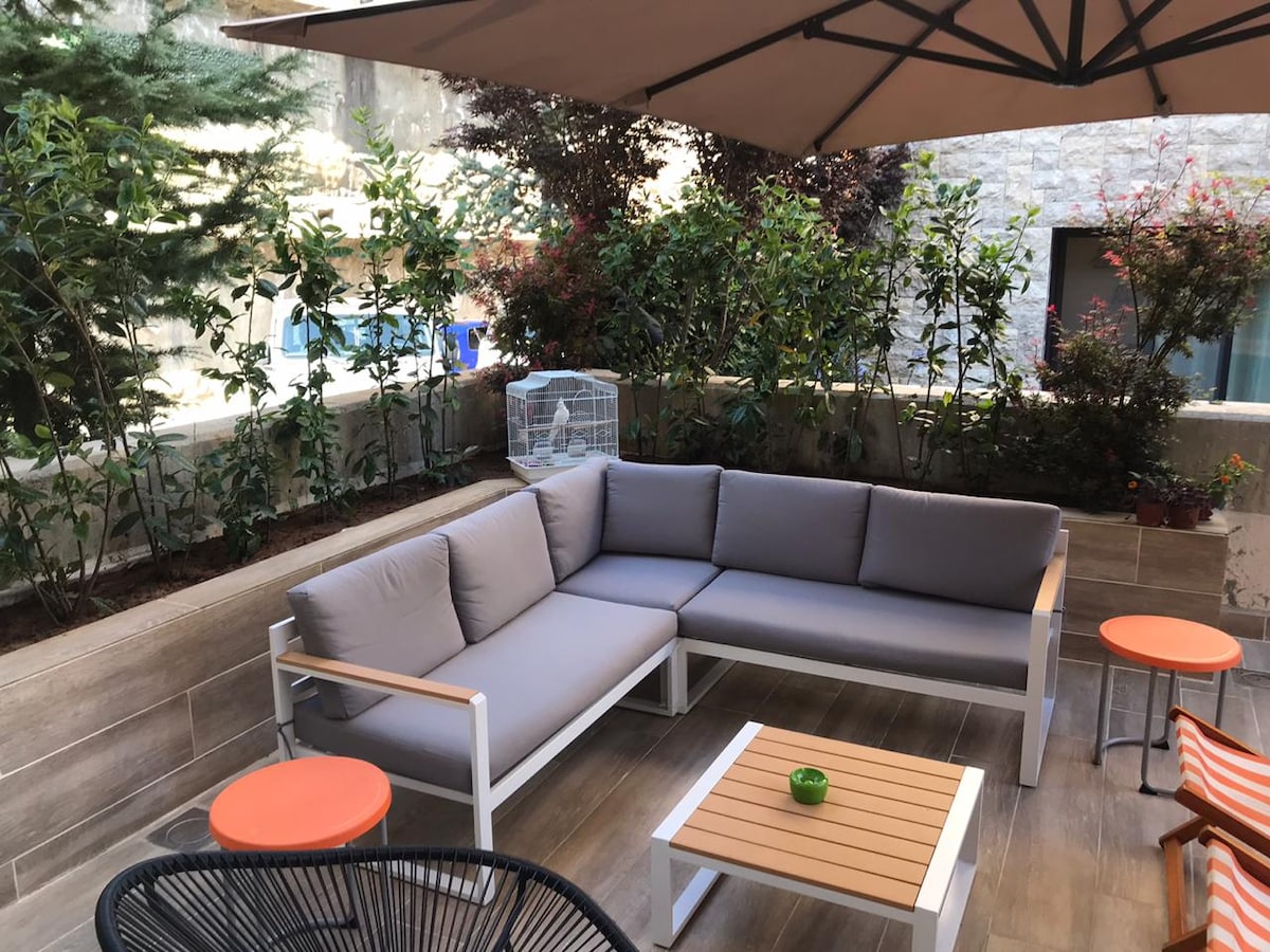 A spacious outdoor terrace is furnished with a gray sectional sofa, a wooden coffee table, and two orange side tables. Lush greenery surrounds the area, providing a comfortable and inviting setting for relaxation. A patterned sunshade offers shade from the sun.