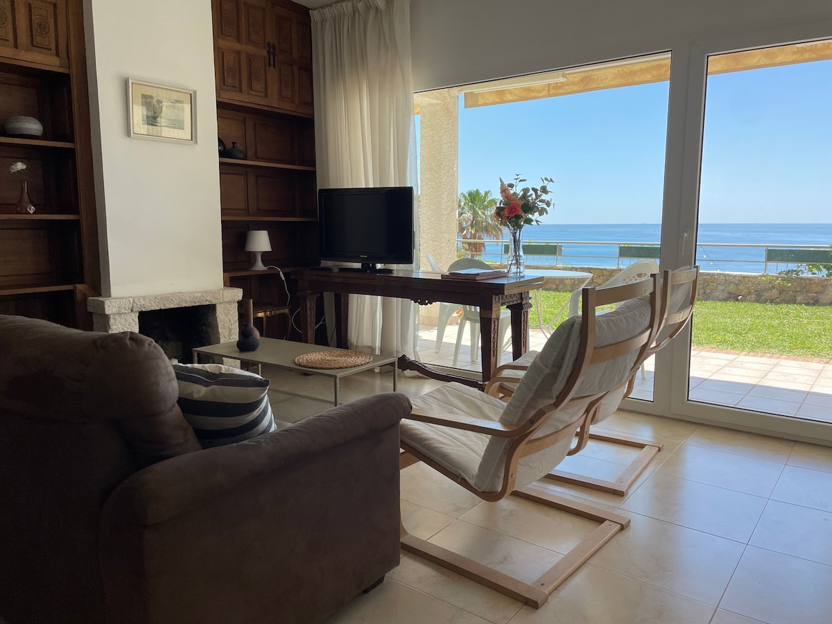 A comfortable living area features a dark sofa and a light wooden chair positioned around a wooden table. Large glass doors open to a private garden with sea views. A TV is placed on a shelf near a stone fireplace, creating a cozy ambiance.