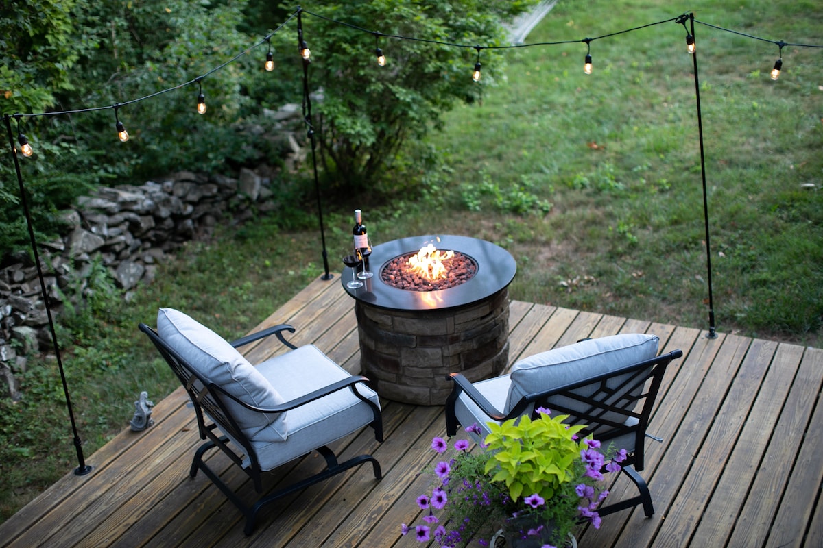A cozy outdoor seating area features two cushioned chairs arranged around a stone fire pit, with a gentle flame flickering in the center. Strand lights hang above, creating a warm ambiance, while colorful flowers add a vibrant touch to the inviting space.