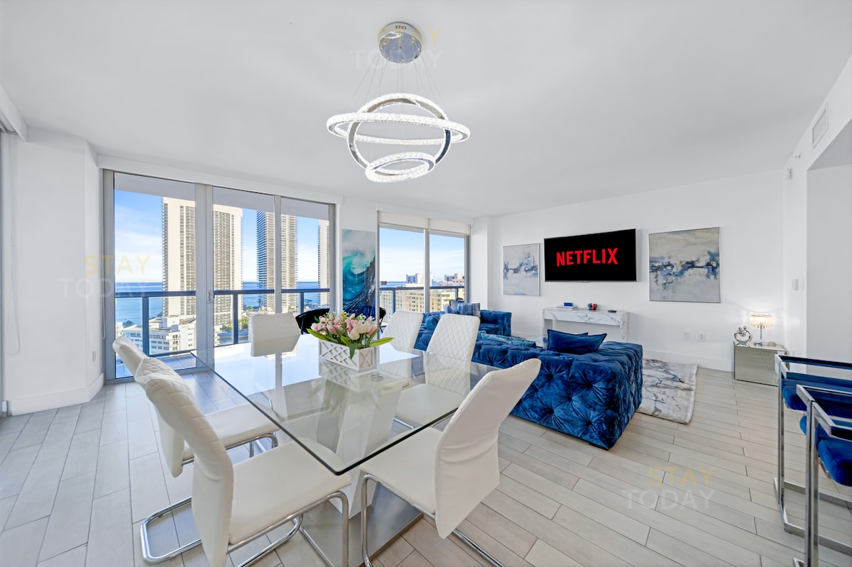 A modern living room features a glass dining table surrounded by six white chairs. A blue velvet sectional sofa offers ample seating, while a large flat-screen TV displays Netflix. Floor-to-ceiling windows provide views of the ocean and city skyline.