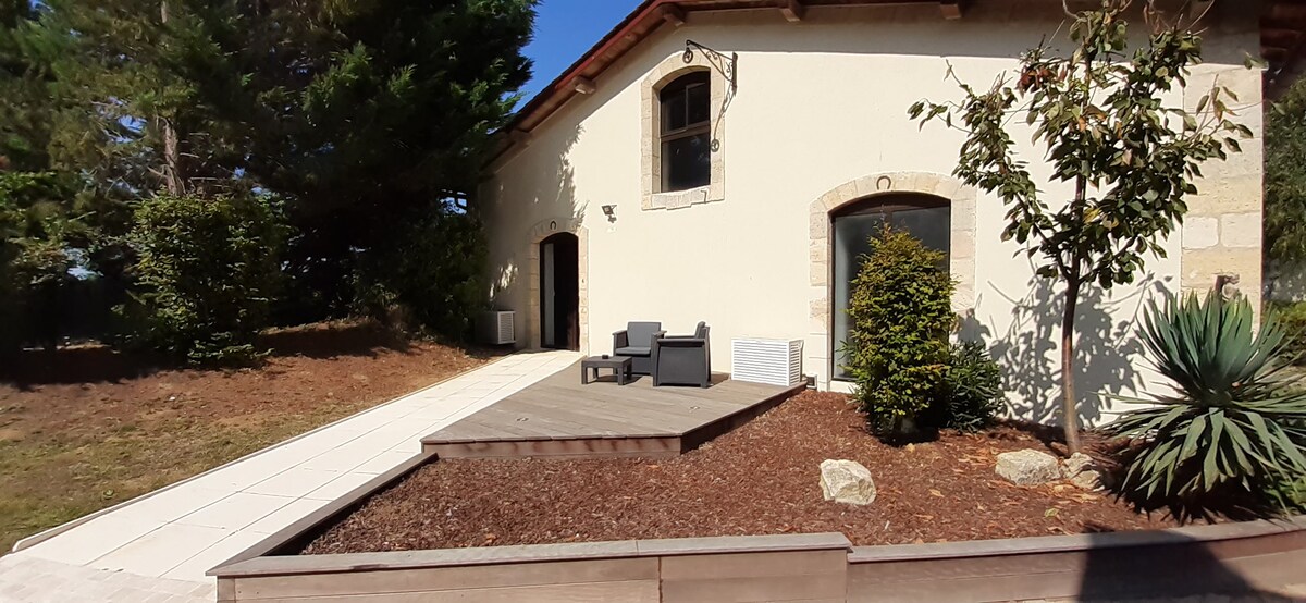 A well-maintained exterior area features a wooden deck with two modern chairs and a small table, surrounded by landscaped greenery. The building's rustic facade, with large windows and natural stone elements, complements the serene outdoor space.