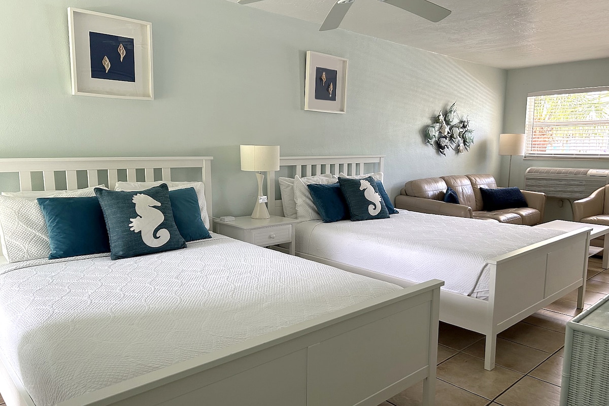 A bright and airy studio features two spacious queen-sized beds adorned with crisp white linens and blue decorative pillows. A small bedside table and lamps are positioned between the beds. A cozy seating area contains a brown couch and a window adorned with light curtains.