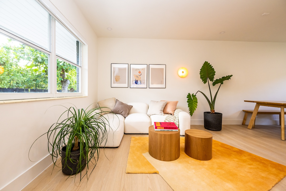 A bright living area features a white sectional sofa adorned with soft pillows. Two wooden side tables sit on a warm-toned rug, accompanied by a tall plant and botanical artwork on the walls. Large windows allow natural light to brighten the space.