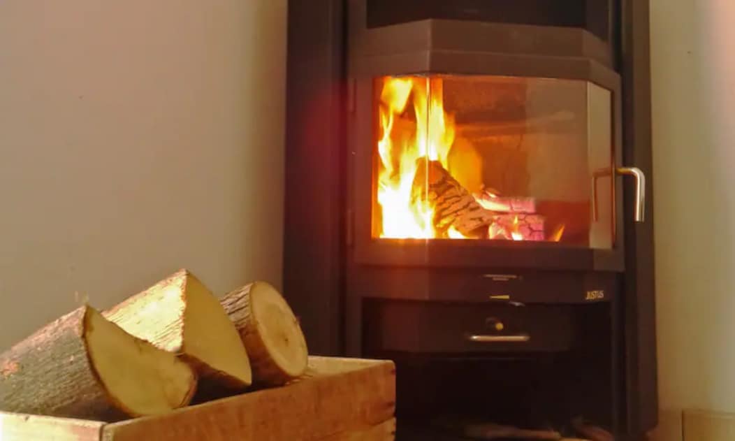 A modern wood-burning stove is positioned against a wall, with flames visible through the glass front. A stack of firewood is placed in a wooden crate nearby, creating a warm and inviting focal point.